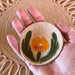 A Globe Mallow Sgraffito Tea Rest by Tough Kitty Designs held in a hand, displaying the unique hand-carved orange flower and green leaves design, measuring 3 inches in diameter.