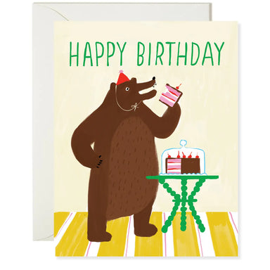 A cheerful birthday card showcasing a bear wearing a party hat, holding a slice of cake in one hand with a cake display on a table. The text says 'Happy Birthday.' From the Karen Schipper Card Collection.