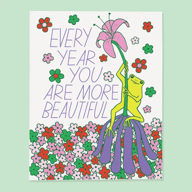 Colorful greeting card illustrating a frog holding a flower, surrounded by bright flowers, and the phrase 'EVERY YEAR YOU ARE MORE BEAUTIFUL', designed by The Good Twin as part of 'The Good Twin Card Collection'.