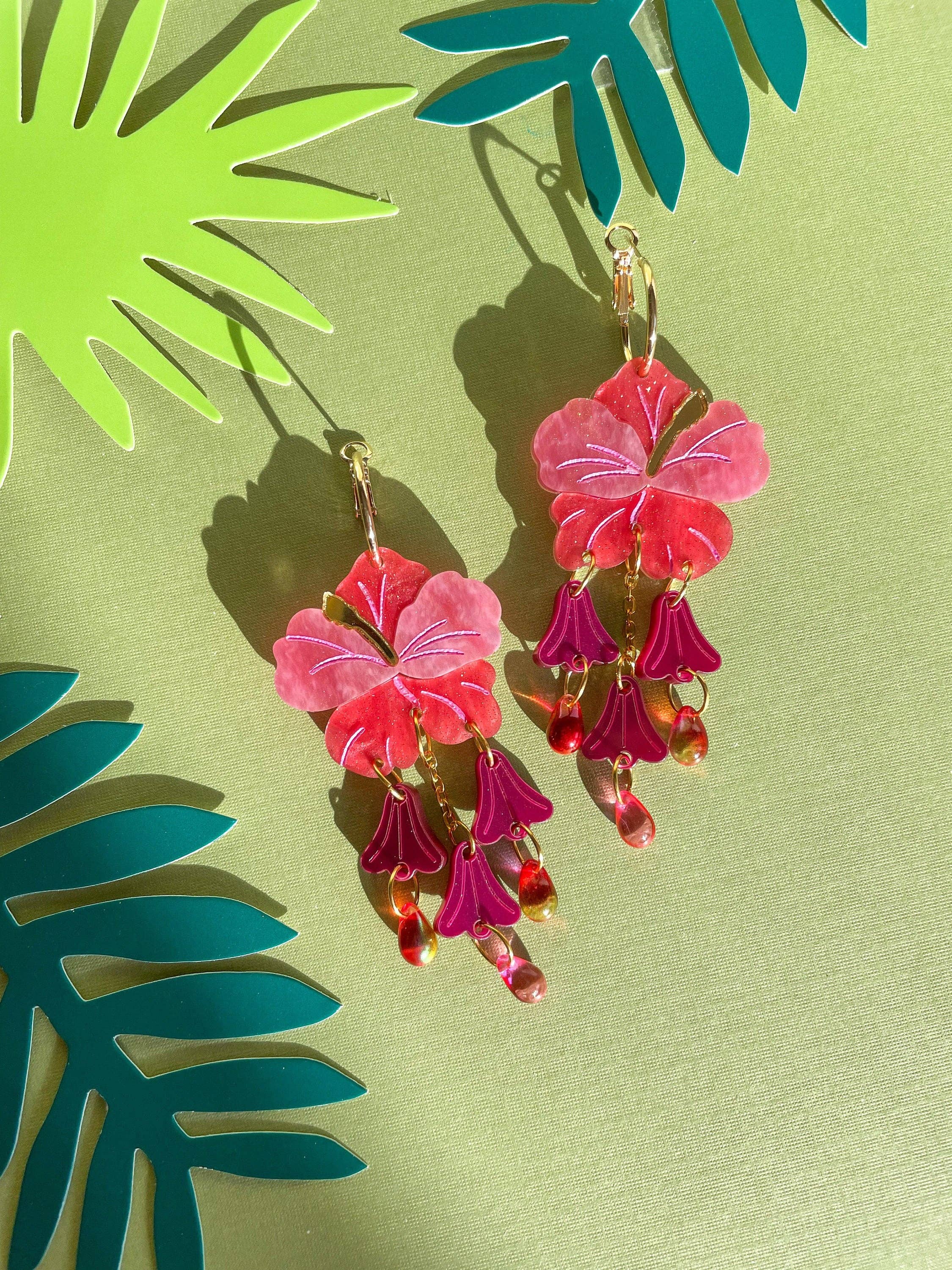 Vibrant Hibiscus Dangle earrings by Sunnie Creative, featuring pink and purple floral designs with dangling accents, displayed against a green backdrop with tropical leaves.