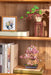A beautifully arranged shelf displaying the Sakura Bonsai Modern Wooden Puzzle by Hands Craft with a cherry blossom bonsai puzzle next to books and a framed artwork.