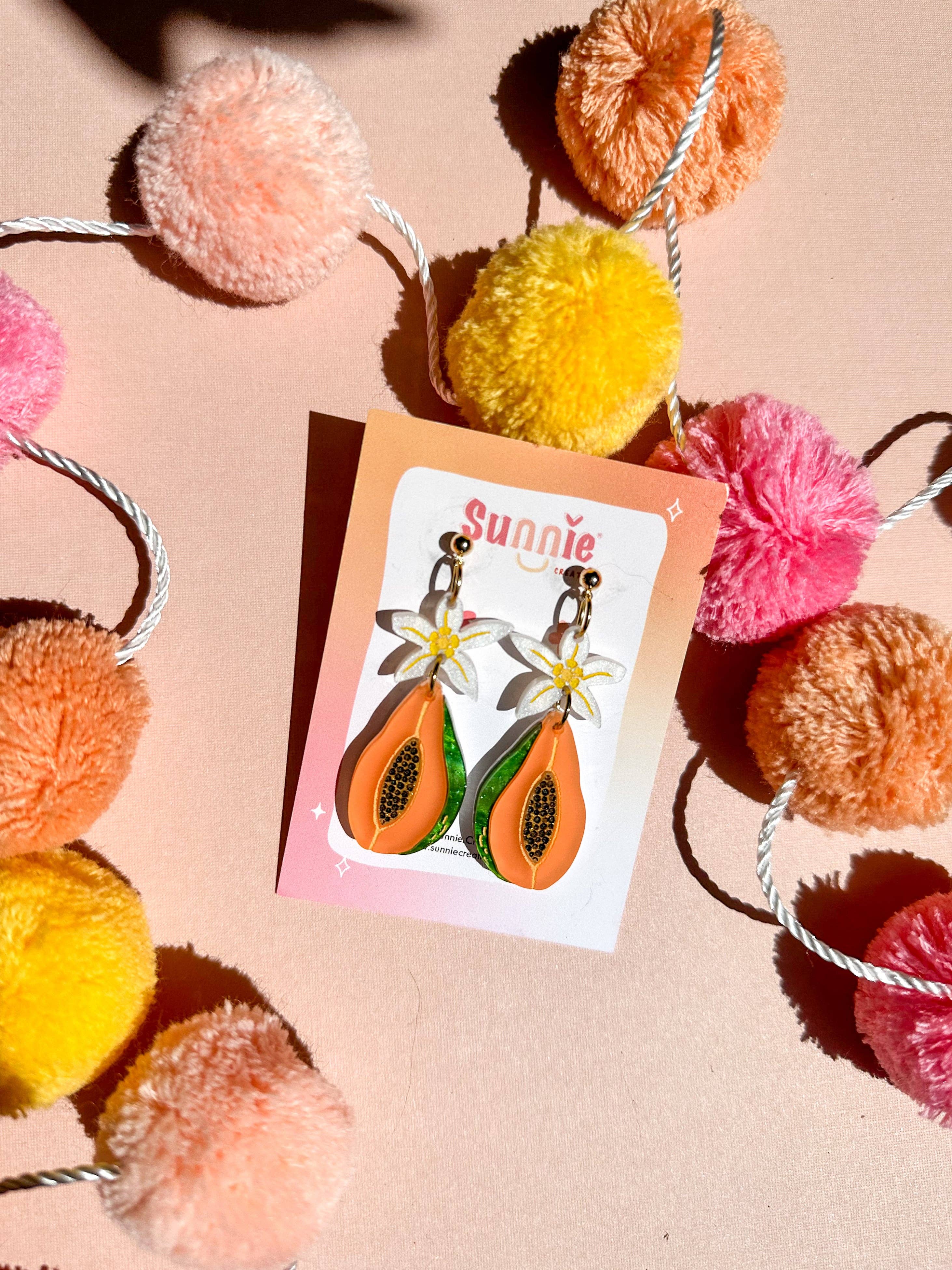 Papaya Dangle earrings from Sunnie Creative displayed with branding on a colorful card, surrounded by playful pom-poms, emphasizing their unique handmade quality.