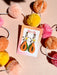 Papaya Dangle earrings from Sunnie Creative displayed with branding on a colorful card, surrounded by playful pom-poms, emphasizing their unique handmade quality.