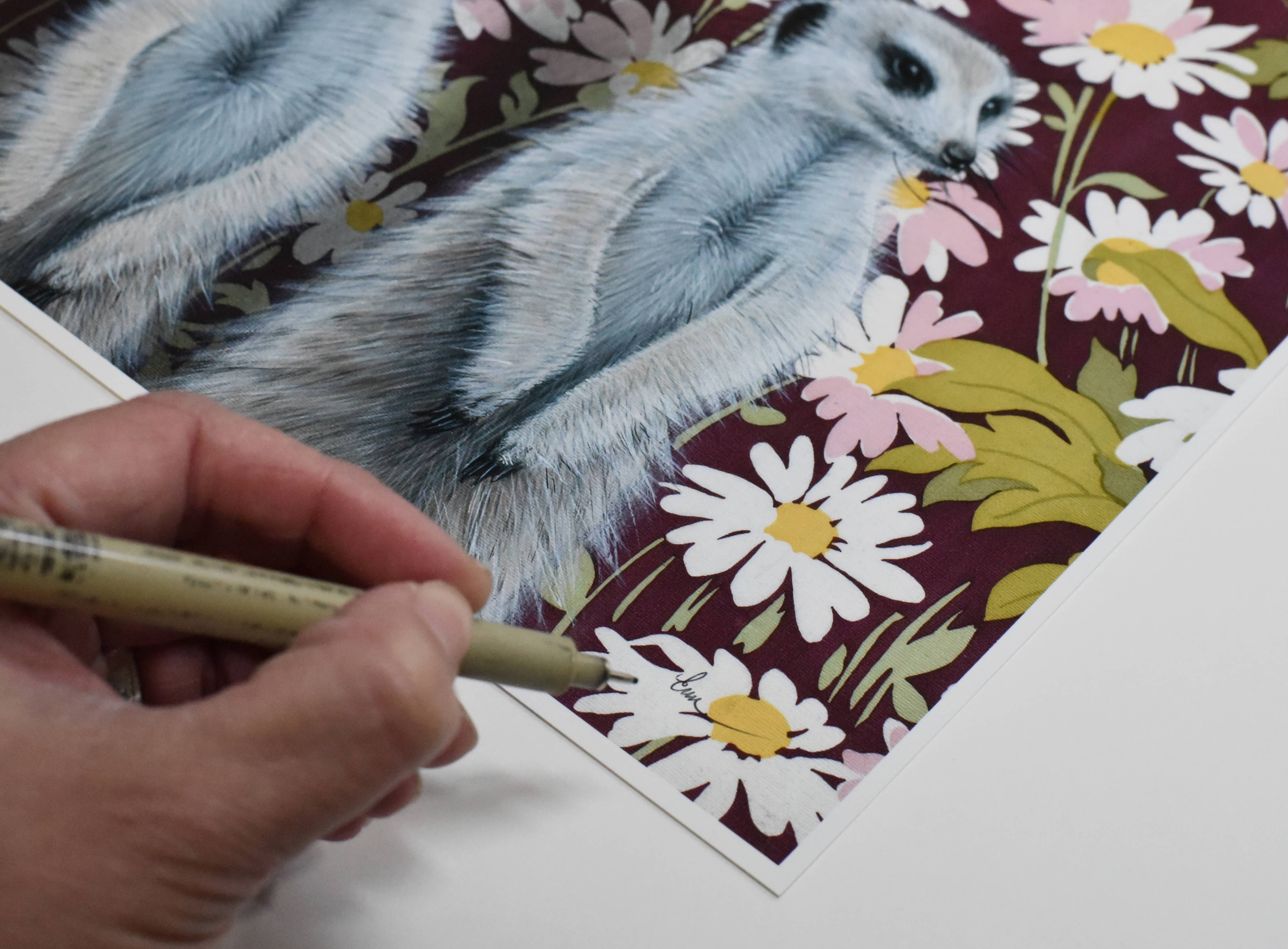 Emily Uchytil hand-signing the meerkats fine art print, capturing the artist's personal touch on the floral-themed artwork.