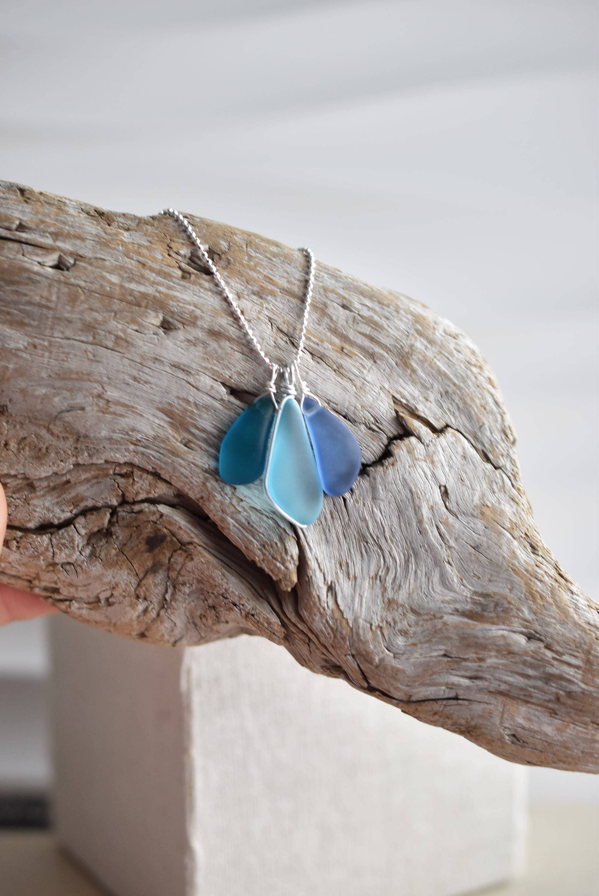 Handcrafted Sterling Silver Recycled Glass Necklace by Whitelotus Design, featuring three frosted glass drops in layered shades of blue, elegantly displayed on a natural wood background.