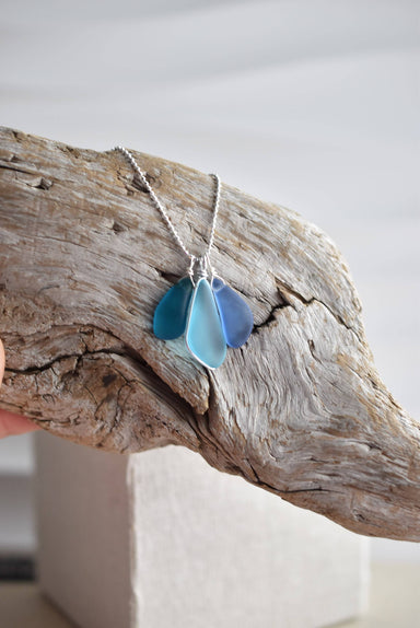 Handcrafted Sterling Silver Recycled Glass Necklace by Whitelotus Design, featuring three frosted glass drops in layered shades of blue, elegantly displayed on a natural wood background.