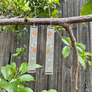Honeysuckle Stained Glass Resin Earrings by Wild Lupine Folkcraft hanging on a natural branch, highlighting their unique design and vibrant colors against a rustic wooden backdrop.