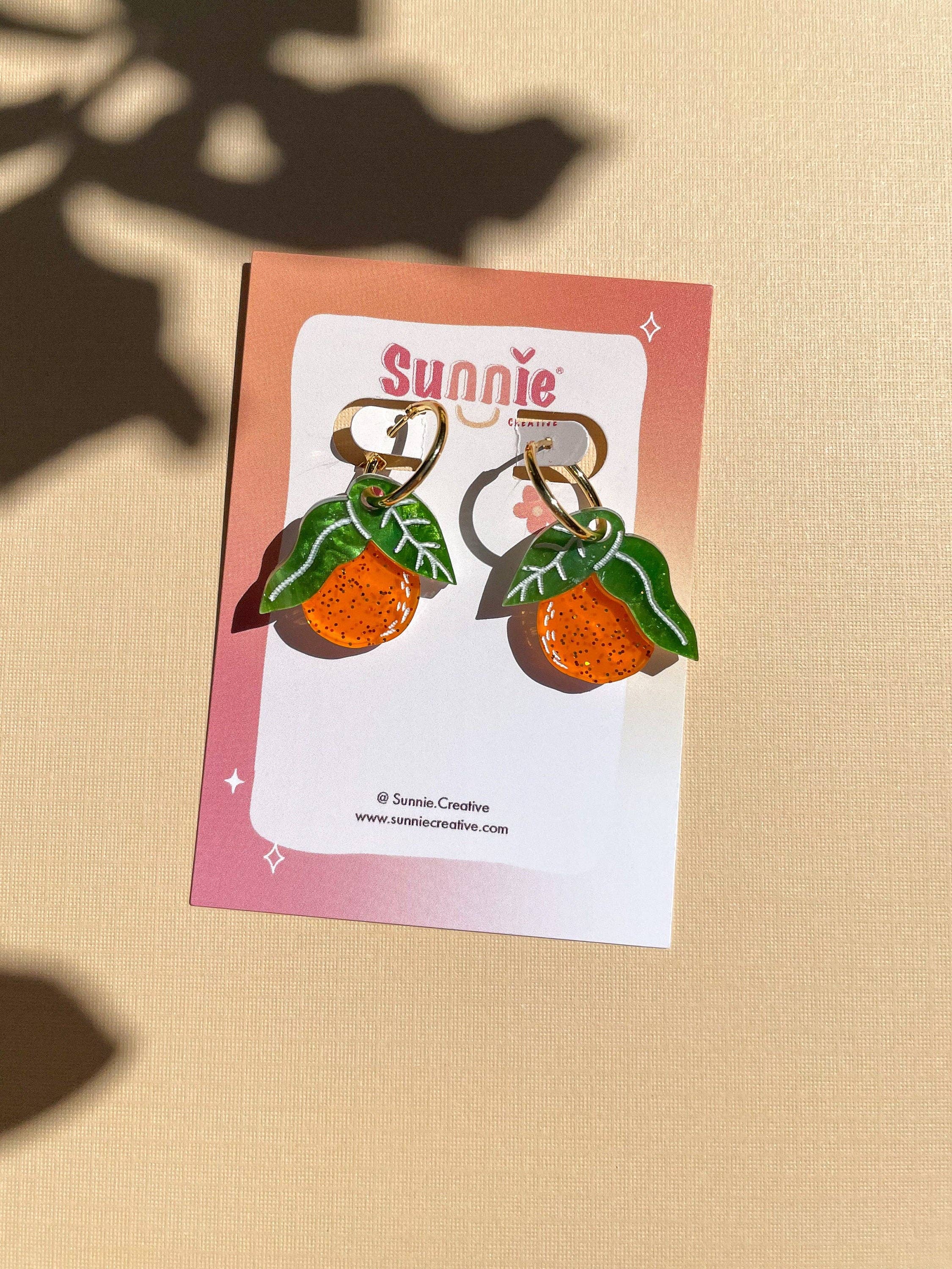 Mini Orange Dangle earrings by Sunnie Creative showcased on a branded card, highlighting their unique orange fruit design and hypoallergenic features, set on a light pastel background.