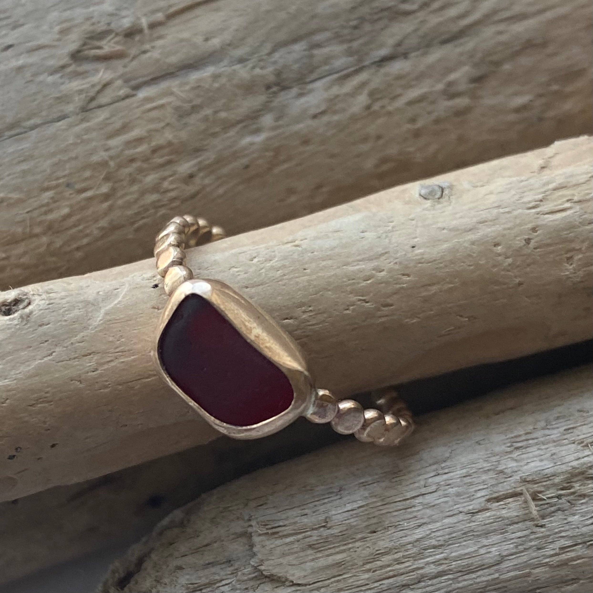 The Repetition Band | Sea Glass Ring by Kate Samson Design featuring a rich red sea glass setting on a textured 14k gold band resting on driftwood.