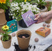 A person preparing to plant a Bespoke Blooms seed ball while holding The Alchemist's Garden kit by Modern Sprout, surrounded by blooming plants in a garden setting.