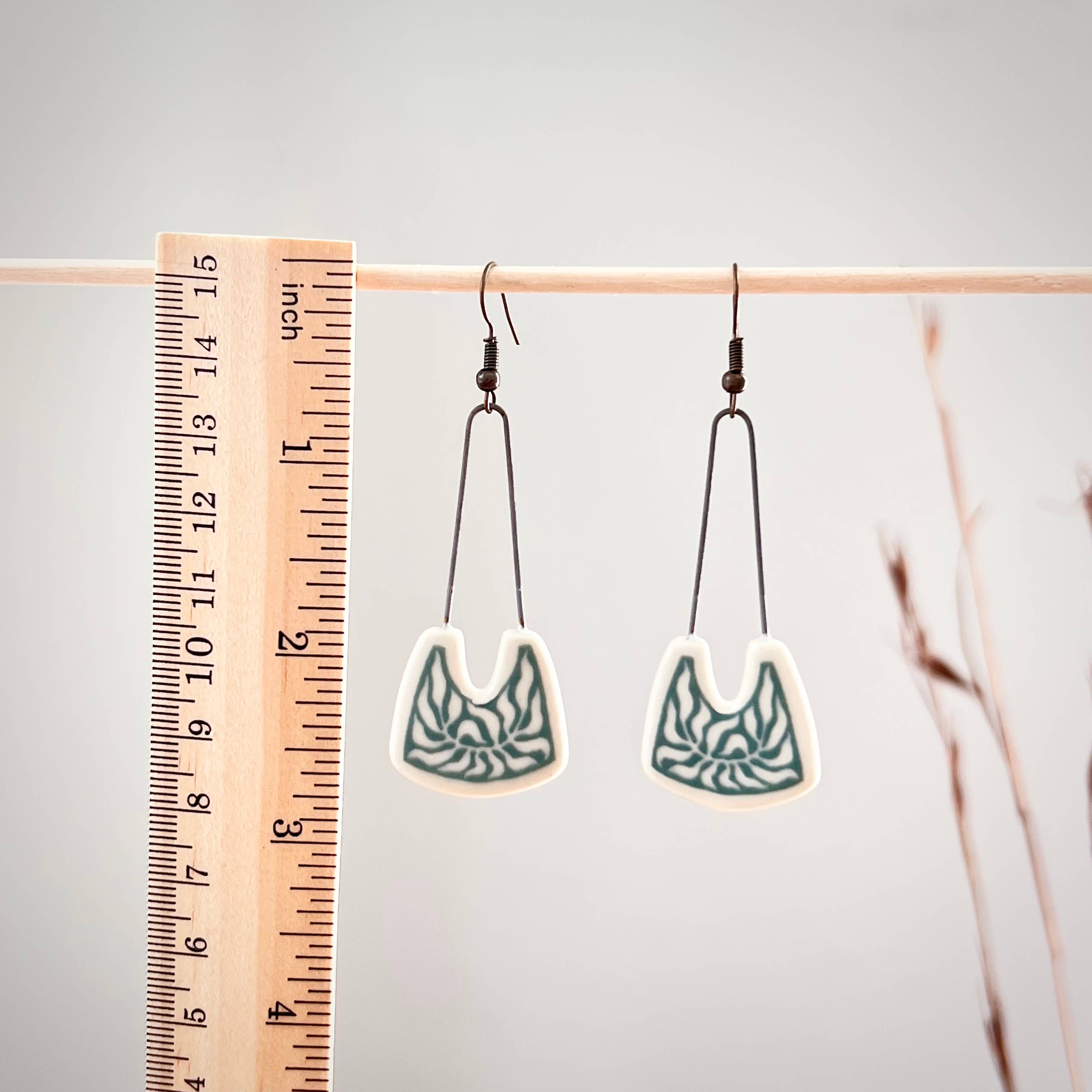 Teal Negative Space Leafy Earrings by The Artifact House hanging on a wooden ruler to show size, featuring unique hand-drawn leaf designs.