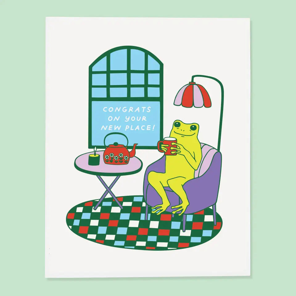 Playful housewarming card with a yellow frog relaxing in an armchair next to a tea set, featuring the text 'CONGRATS ON YOUR NEW PLACE!' from The Good Twin Card Collection, designed by The Good Twin.