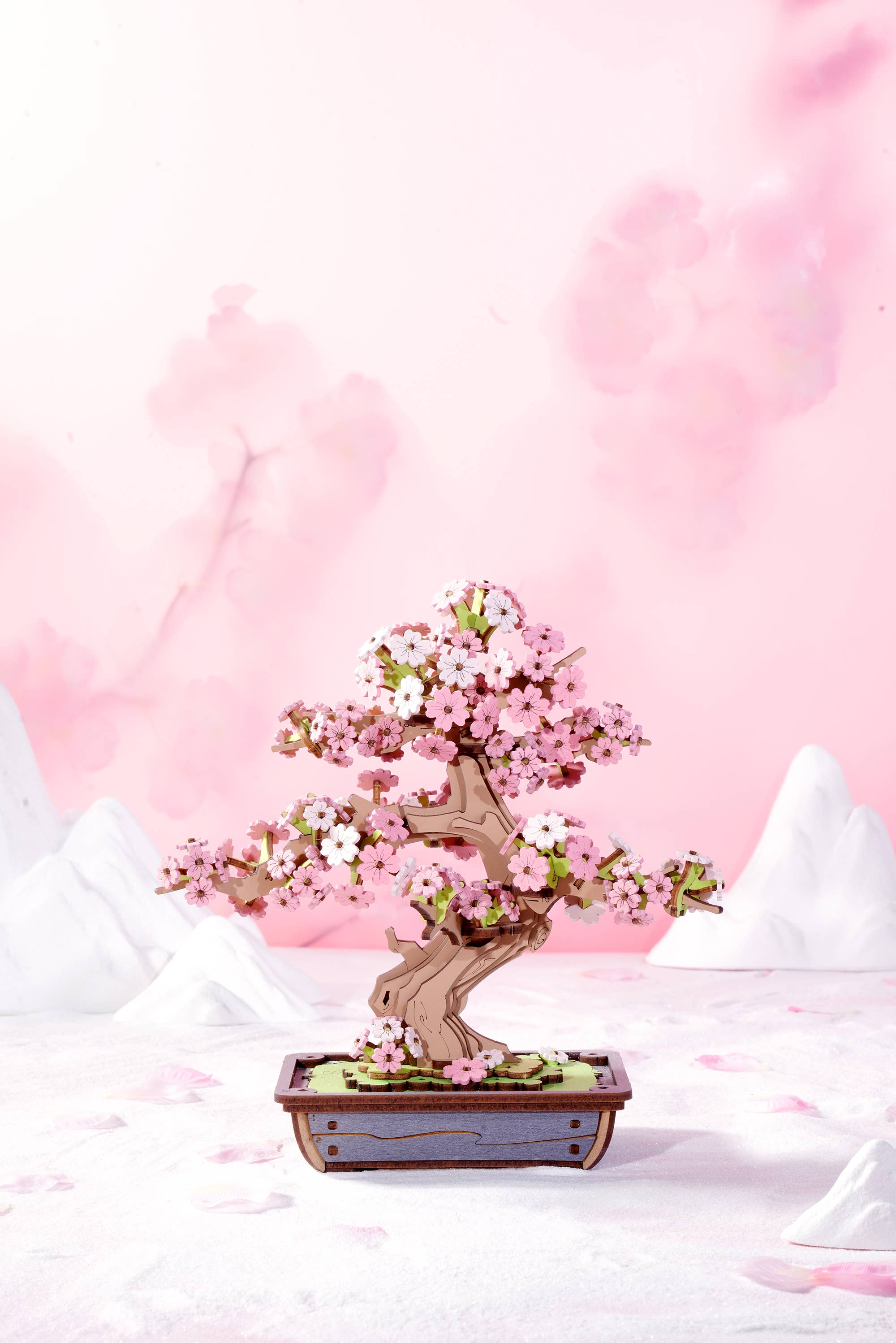 Sakura Bonsai Modern Wooden Puzzle by Hands Craft, presenting an elegant cherry blossom bonsai tree puzzle in pink hues, surrounded by a soft pink backdrop and snow-capped mountains.