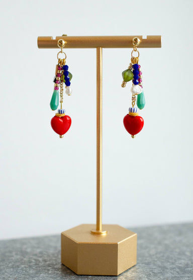 Heart Charm Earrings by Jill Makes showcased on a gold earring stand with a loose cluster of colorful beads and heart charms, set against a blurred background.