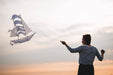 A woman flying a white and blue Sailing Ship Kite by Haptic Lab against a sunset background, illustrating its functionality and beauty in action.