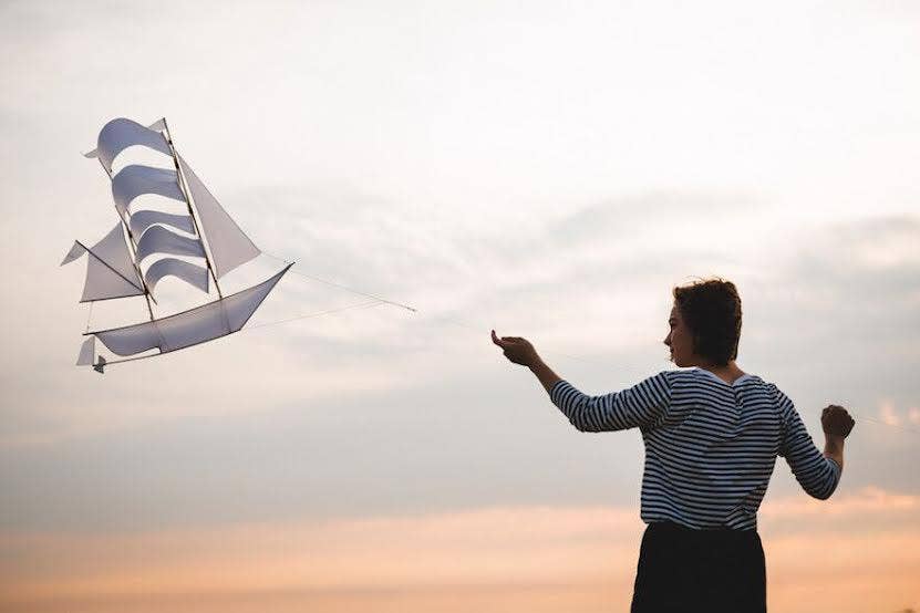 A woman flying a white and blue Sailing Ship Kite by Haptic Lab against a sunset background, illustrating its functionality and beauty in action.