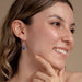Young woman showcasing petite seed earrings by Mosaico Jewels, smiling while highlighting the earrings' colorful design against a neutral backdrop.