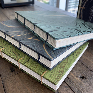 A stack of elegant handmade journals with unique hand-marbled covers, featuring Coptic-style binding, by The Casual Bookbinder. Each journal showcases a distinct marbled pattern in shades of green, blue, and black, inviting creativity in writing and sketching.