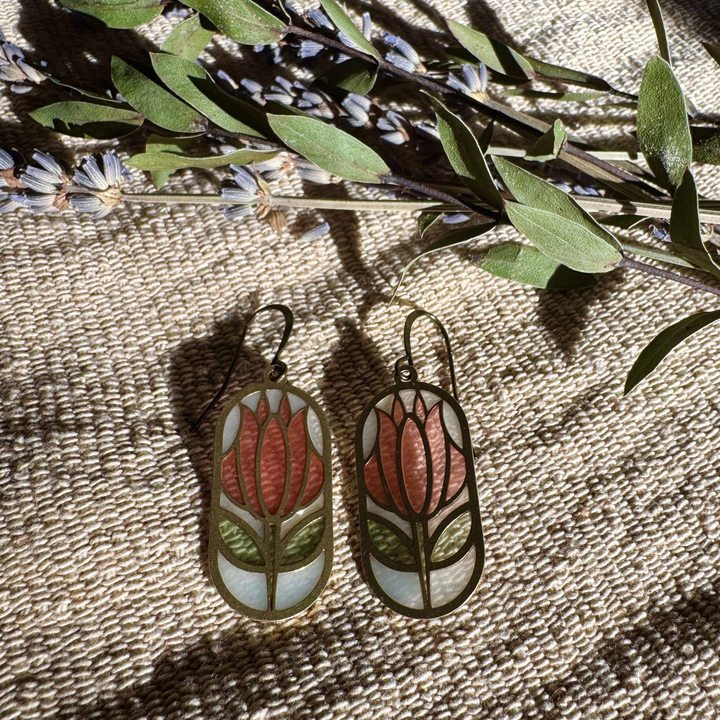 Cloisonné Stained Glass Resin Earrings by Wild Lupine Folkcraft, showcasing a brass geometric botanical design with soft-colored resin stained glass, displayed with natural elements on a textured surface.