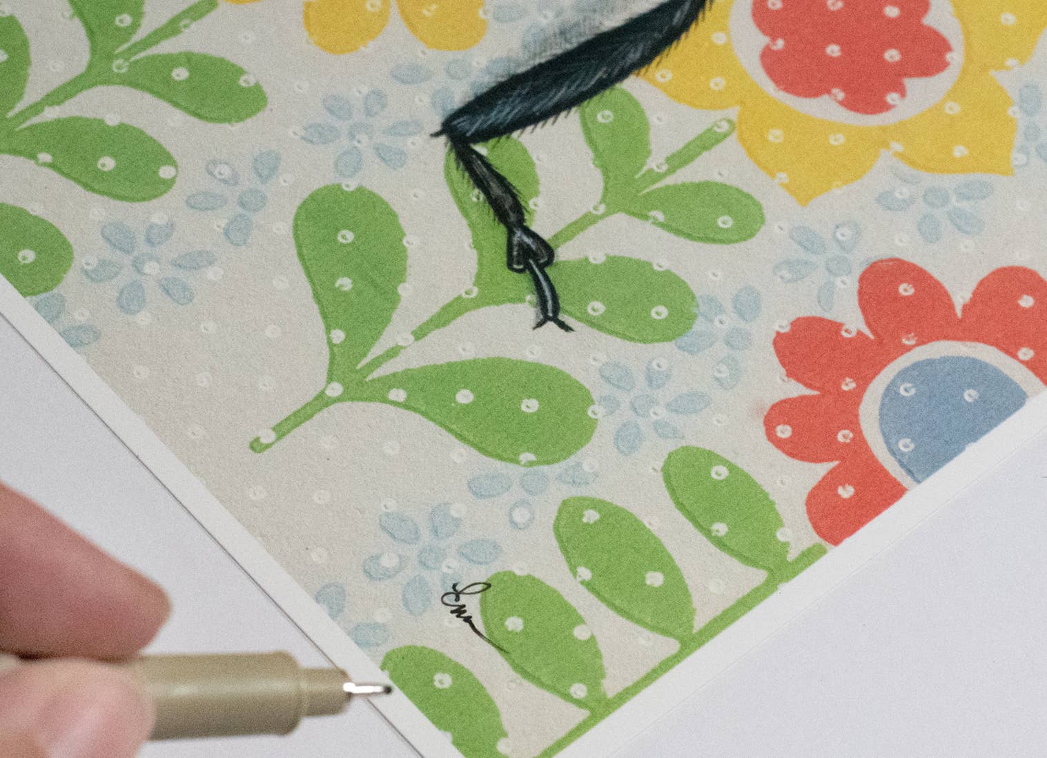 Artist's hand holding a pen next to a ladybug fine art print on patterned paper, showcasing the hand-signed aspect of Ladybug White by Emily Uchytil.