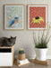 Interior scene showcasing framed art prints titled Bluebird by Emily Uchytil, with a cat sitting beside a plant and decor elements.