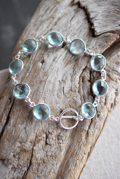 Handmade sterling silver sky blue topaz bracelet by Whitelotus Design featuring faceted gemstones in a modern linked design, showcased on a rustic wooden background.