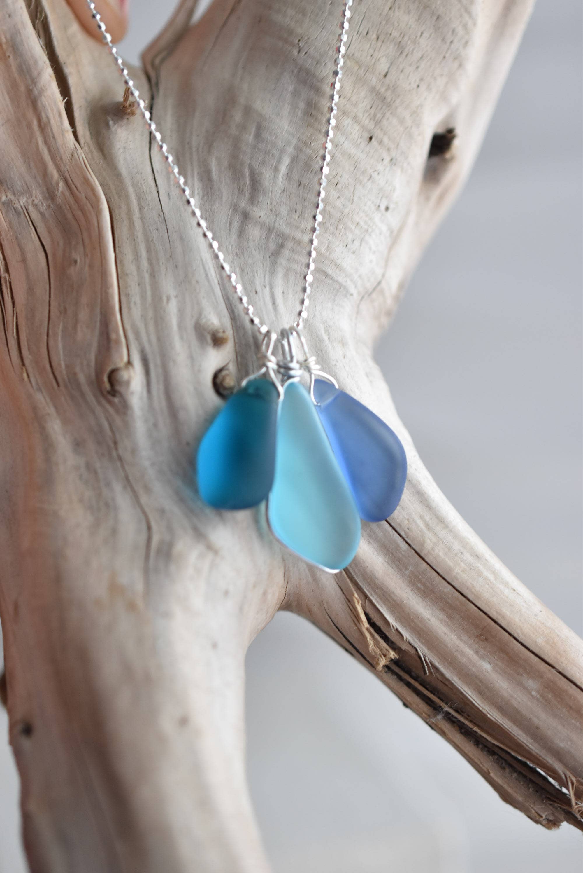 Stylish presentation of the Sterling Silver Recycled Glass Necklace by Whitelotus Design, featuring three smooth, matte-finished blue glass drops against a light wood background.