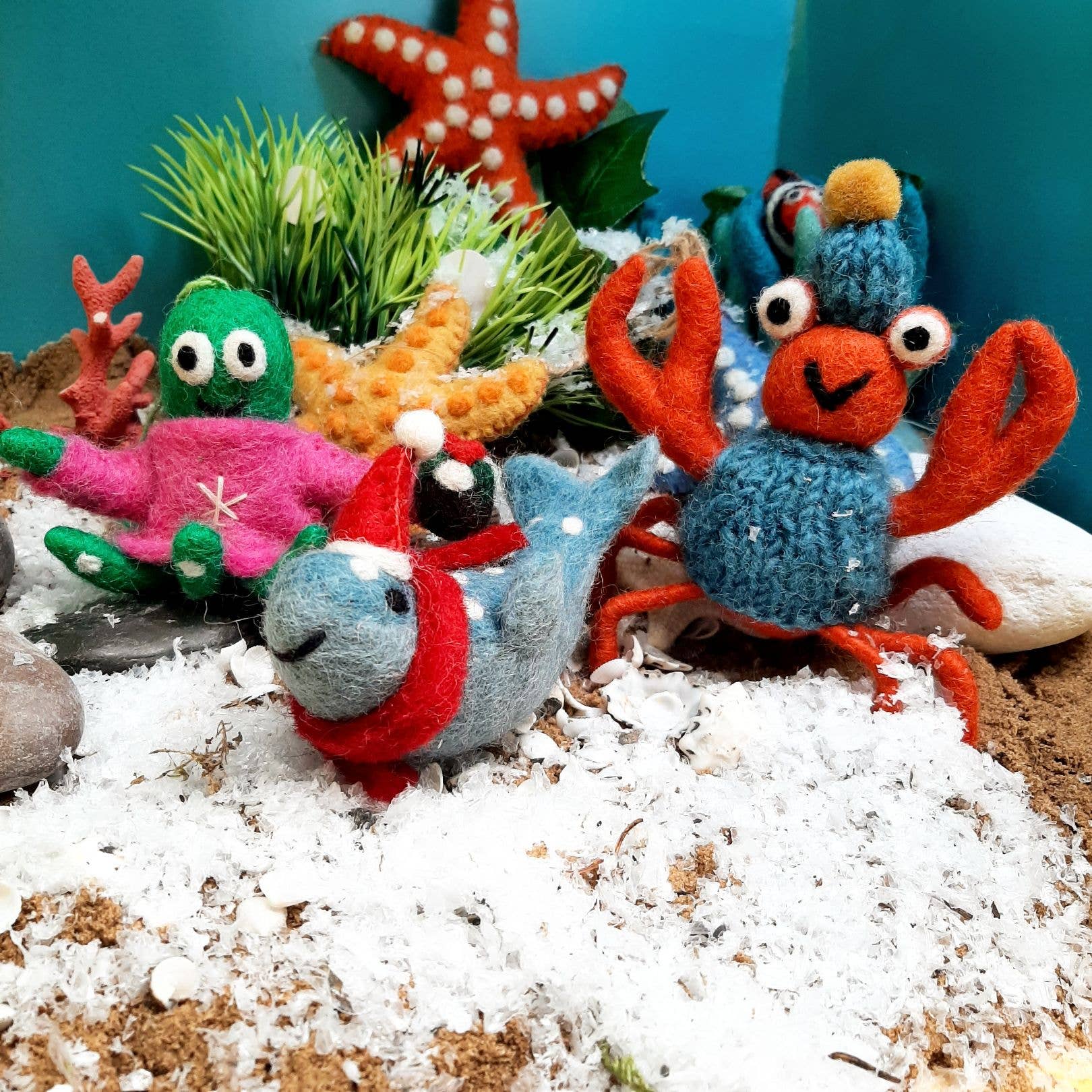 Felt sea creature ornaments on a sandy surface with plants and rocks.