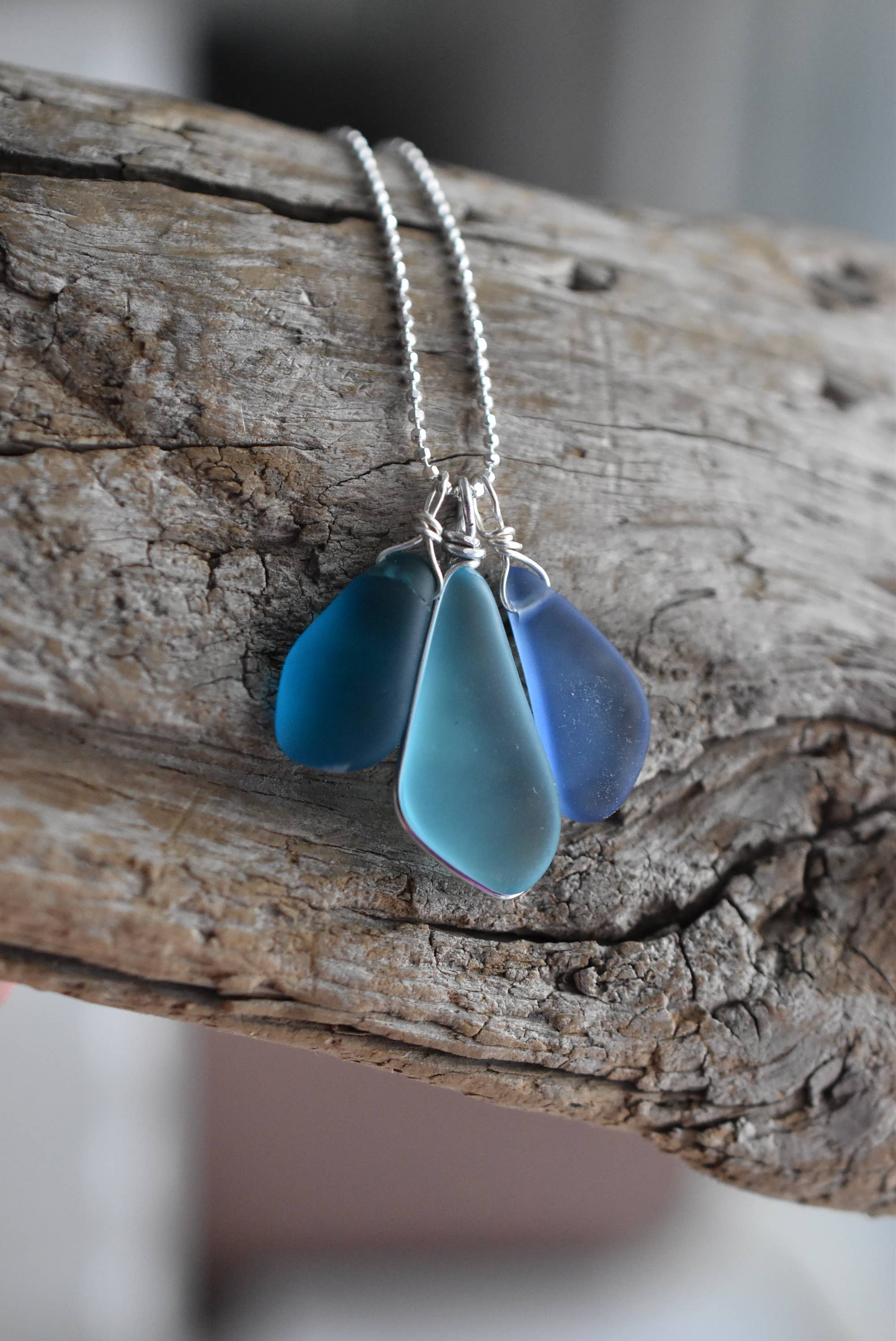 Artistic view of the Sterling Silver Recycled Glass Necklace by Whitelotus Design with three blue frosted glass drops, elegantly clustered on a driftwood display.