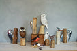 A diverse collection of hand-painted wooden birds, including owls and woodpeckers by Aviologie, arranged on natural wood logs, enhancing indoor decor.