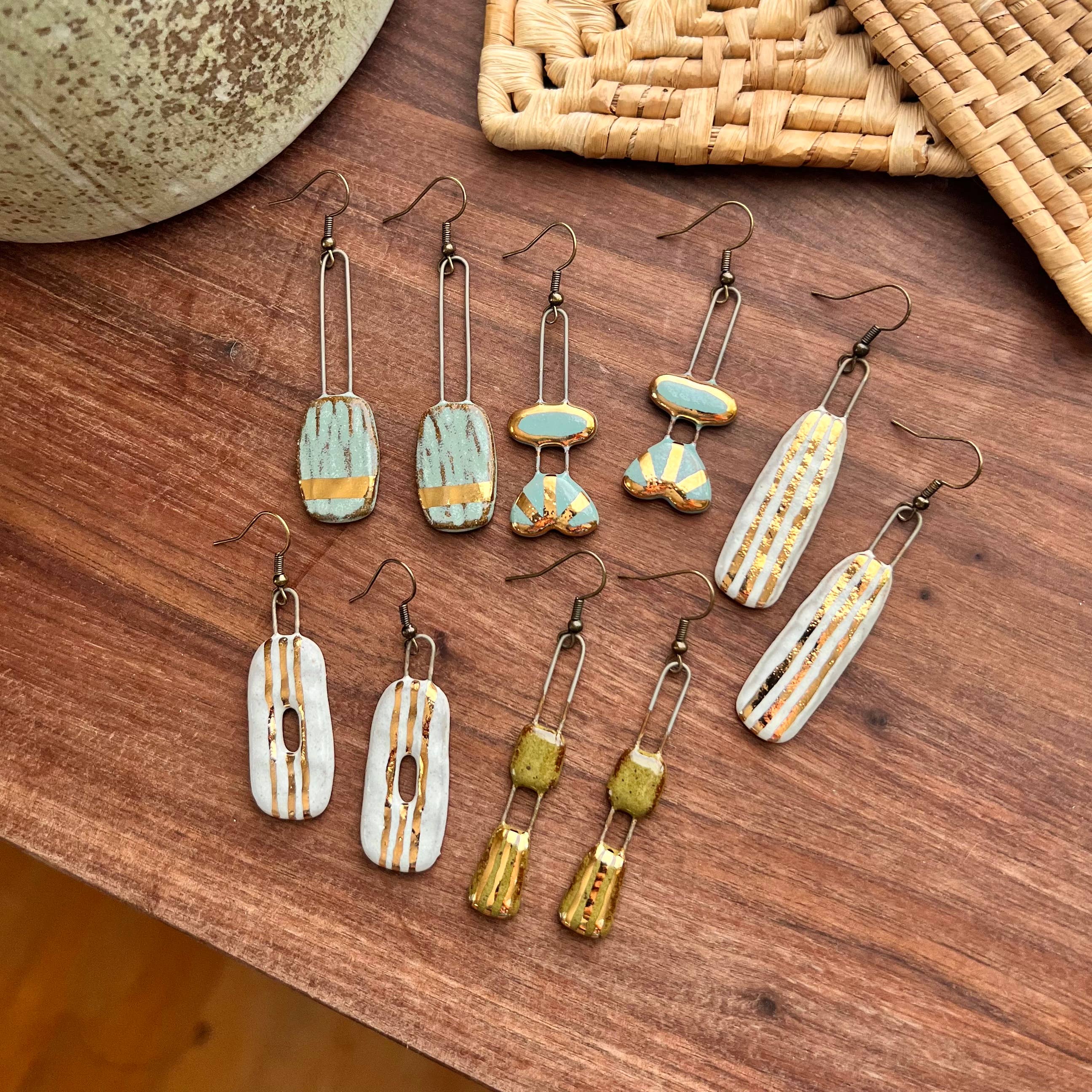 Variety of handmade ceramic earrings including the Speckled Olive Two Piece Gold Lined Earrings by The Artifact House, arranged on a wooden surface with a textured bowl.