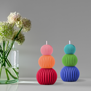 Decorative Fluted Stacked Ball Candle by Made By Humans, featuring a vibrant three-color pattern of pink, orange, and red, displayed next to a glass vase with flowers.