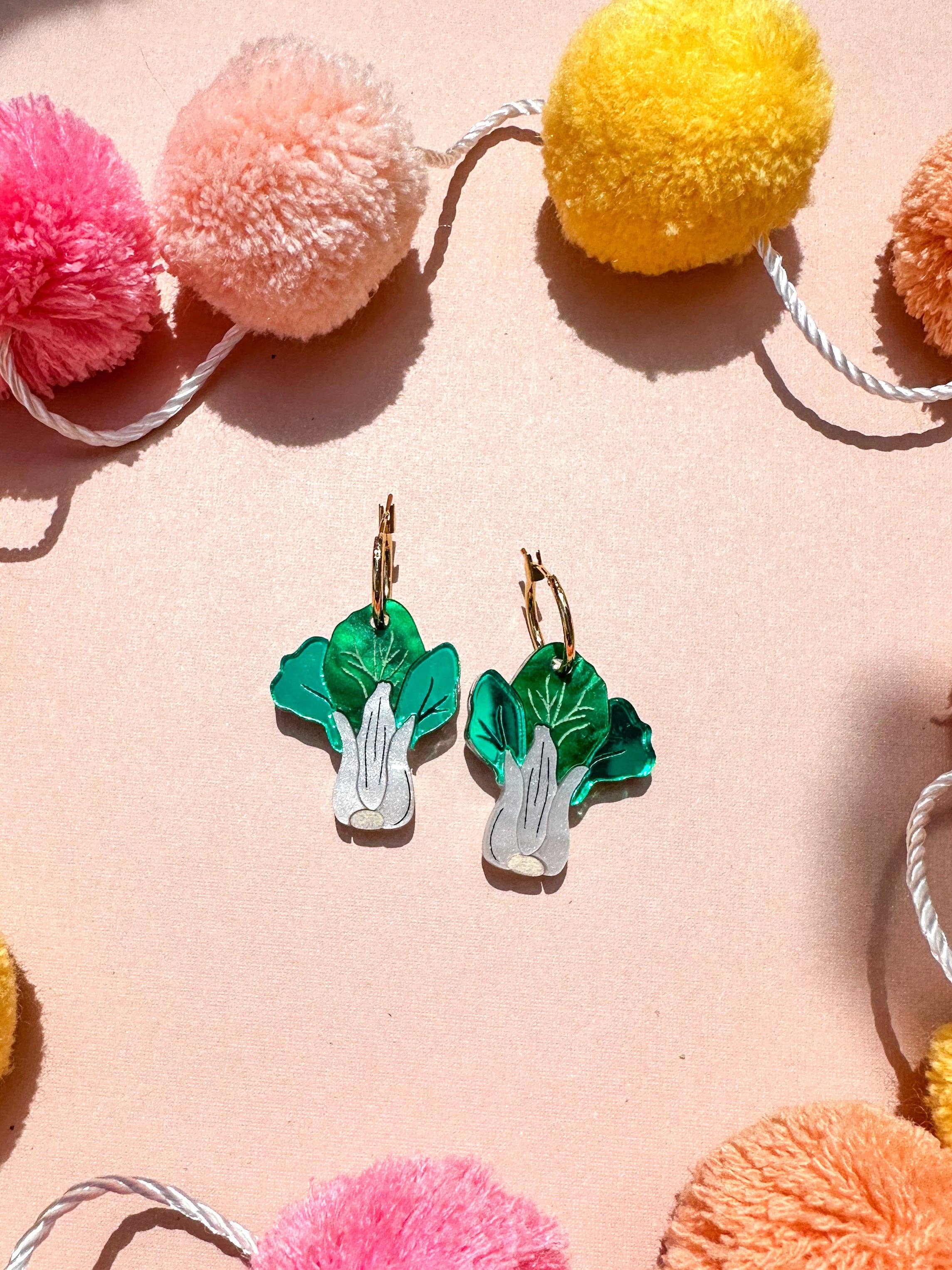 Bok Choy Dangle earrings by Sunnie Creative displayed against a pastel pink background with colorful pom poms, showcasing their vibrant design and handmade details.