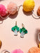Bok Choy Dangle earrings by Sunnie Creative displayed against a pastel pink background with colorful pom poms, showcasing their vibrant design and handmade details.