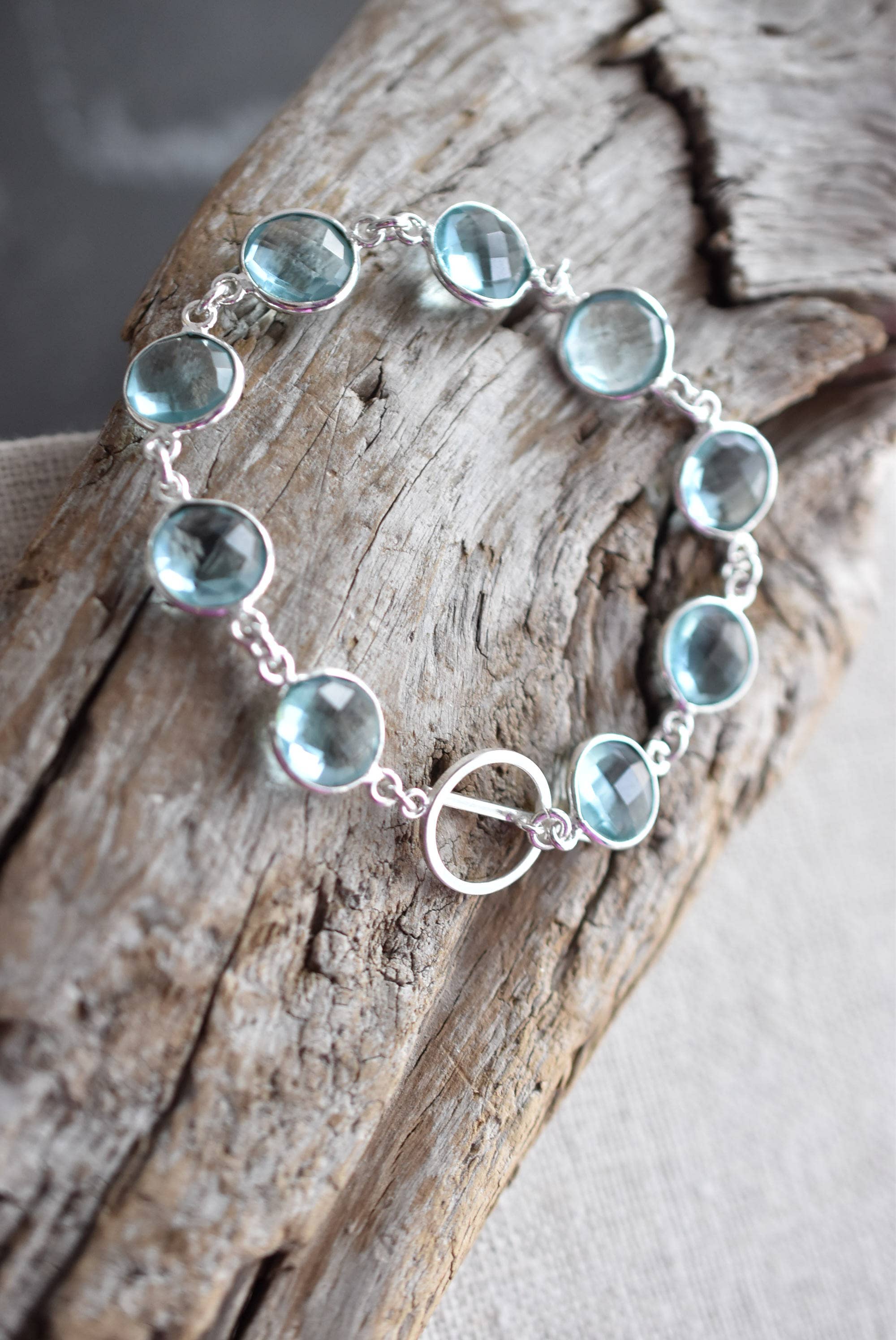 Elegant handmade sterling silver sky blue topaz bracelet by Whitelotus Design, displayed with faceted gemstones, resting on a textured wooden surface.