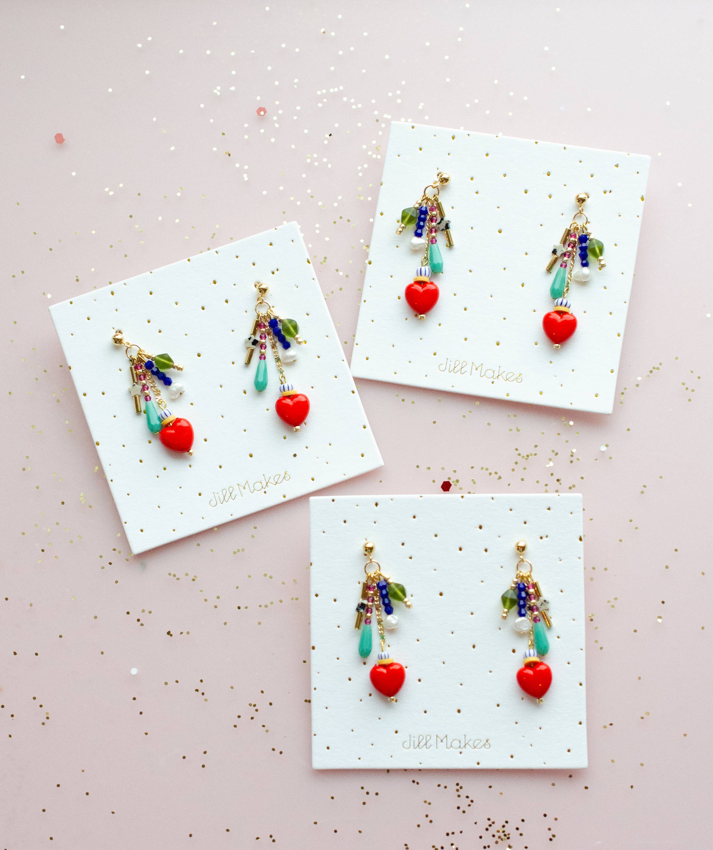 Three pairs of Heart Charm Earrings by Jill Makes arranged on a pink background, featuring vibrant beads and heart designs on decorative backing cards.