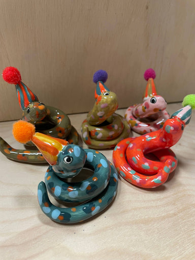 A colorful assortment of Itty Bitty Party Snake Sculptures by Bianca Rae, featuring whimsical designs with party hats topped with pom poms. Each snake is unique in color and style, perfect for cake toppers or charming decor.