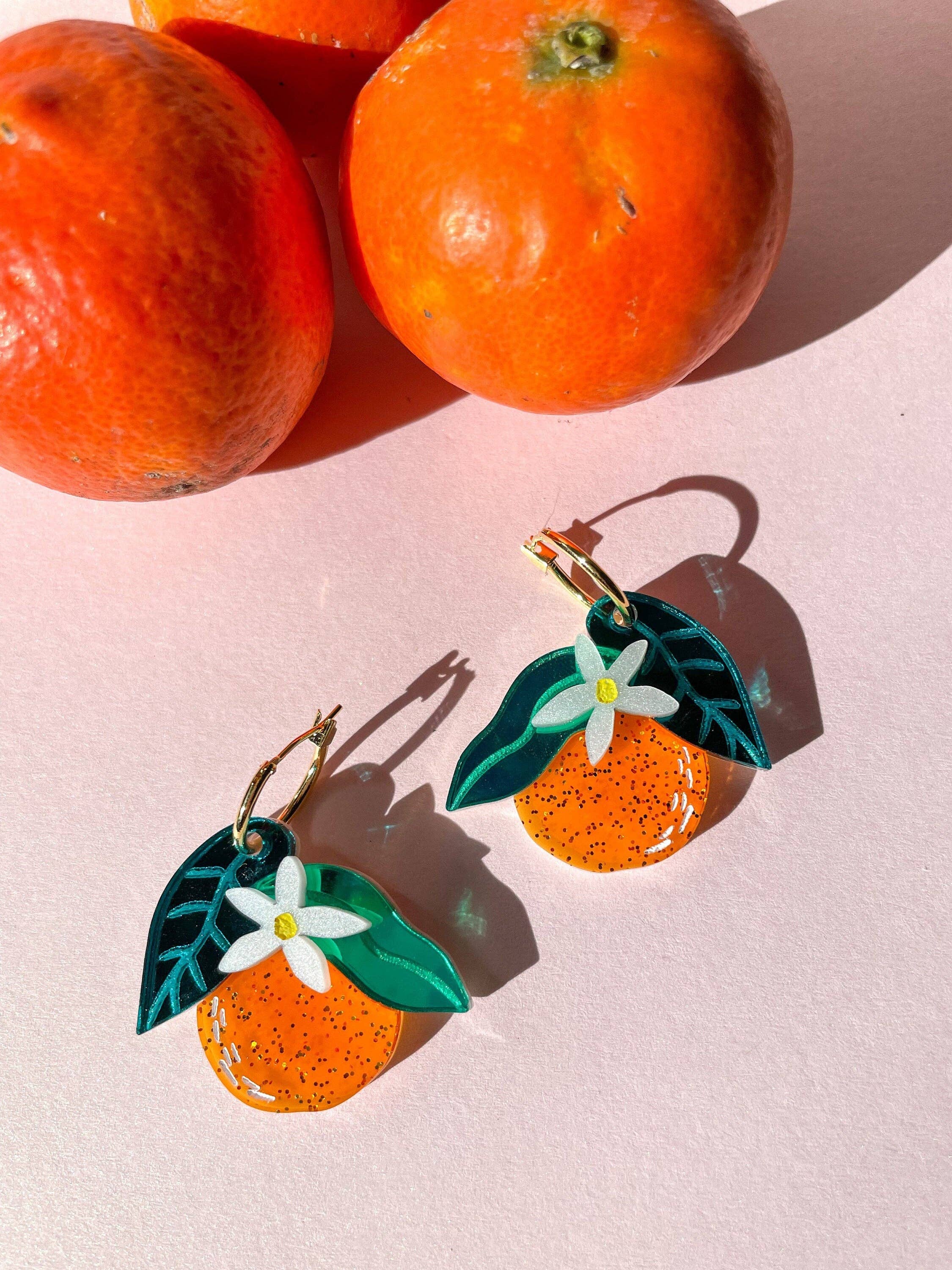 Colorful Orange Dangle earrings displayed with fresh oranges, designed by Sunnie Creative. Unique handmade earrings featuring orange acrylic with leaf and flower accents.