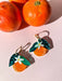 Colorful Orange Dangle earrings displayed with fresh oranges, designed by Sunnie Creative. Unique handmade earrings featuring orange acrylic with leaf and flower accents.