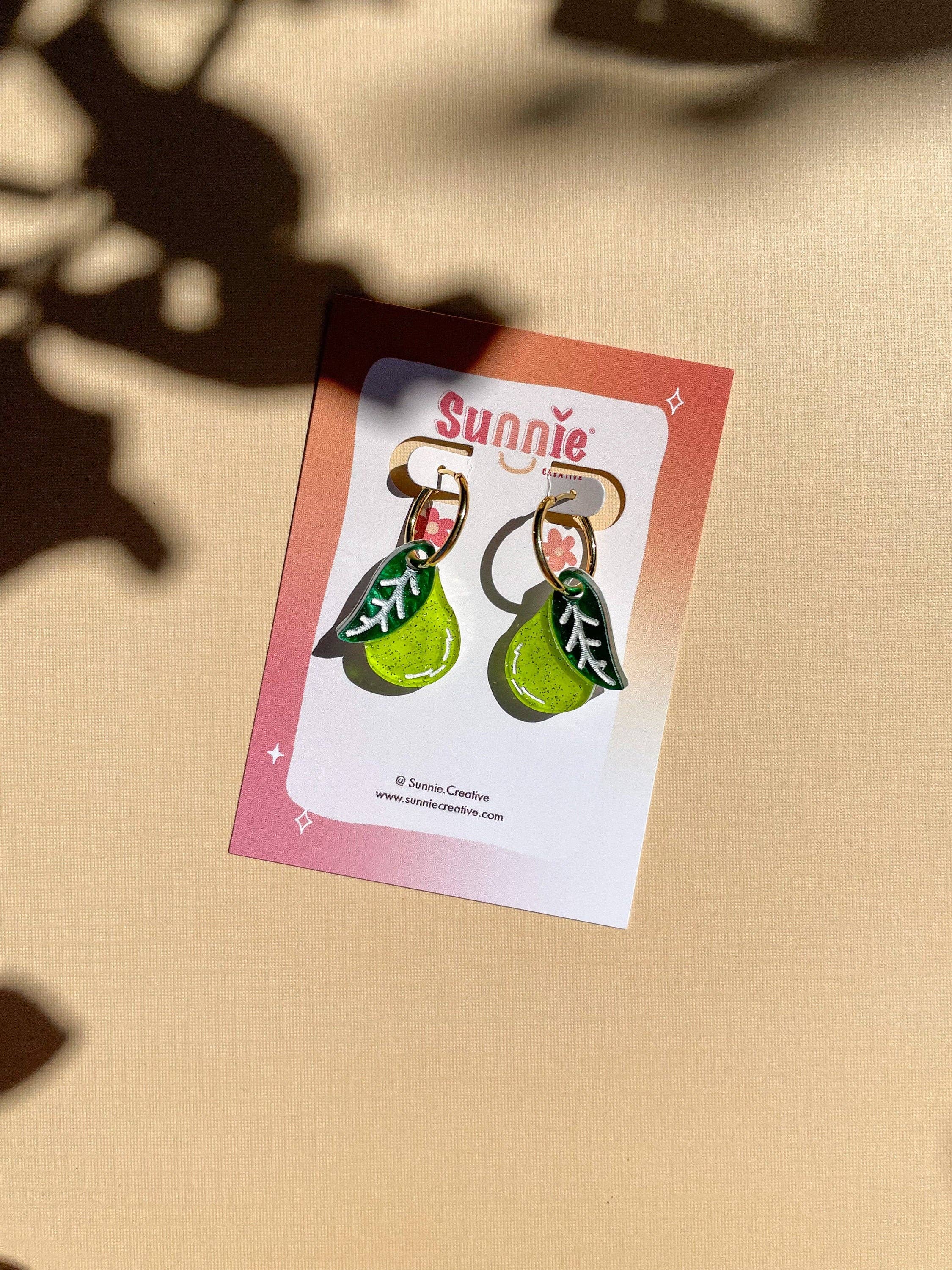 Mini Pear Dangle earrings by Sunnie Creative displayed on a stylish card, featuring a unique design of green acrylic pears with leaf accents, highlighted with natural lighting.