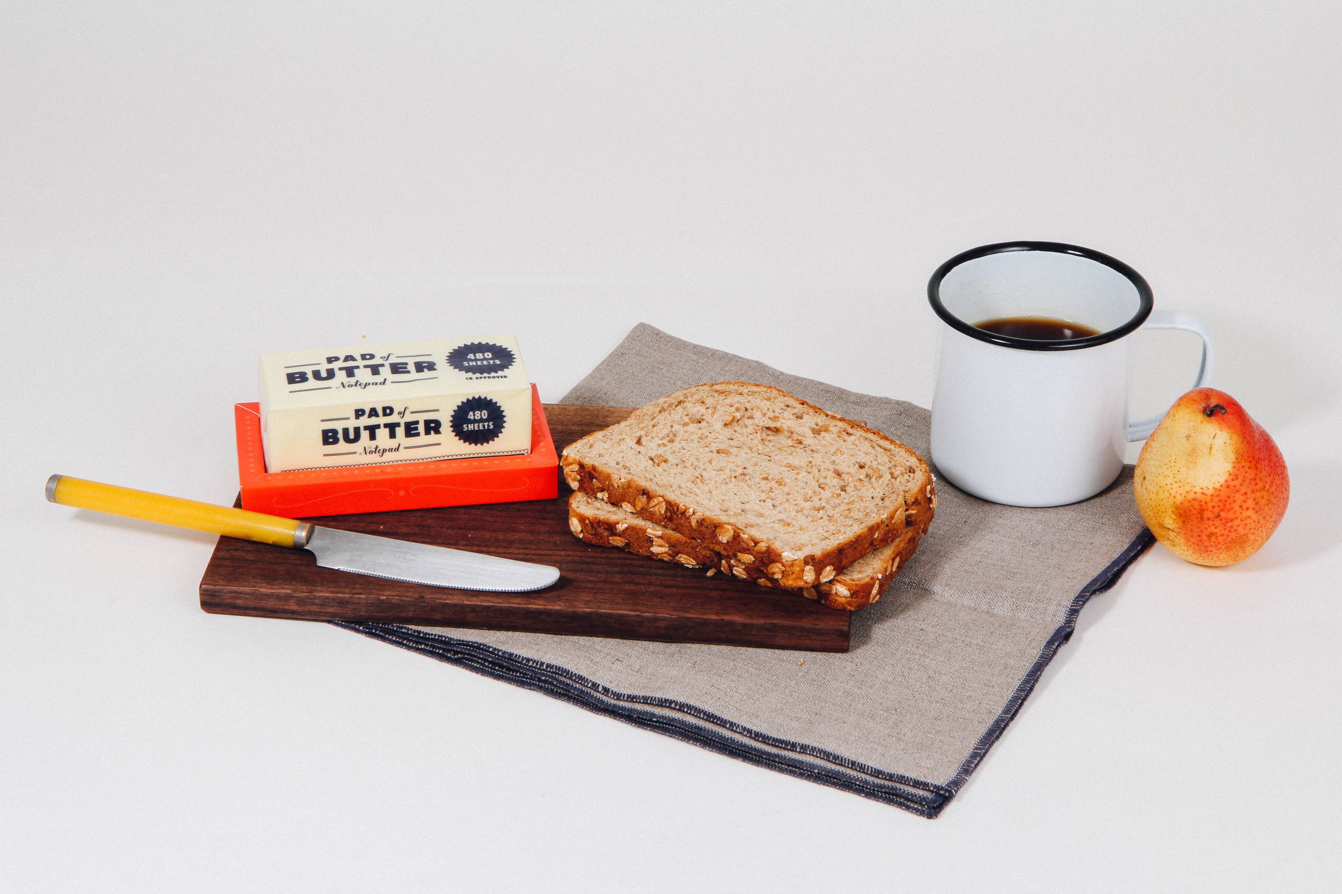 Lifestyle image of the Pad of Butter notepad by Chronicle Books displayed with a slice of bread, coffee, and apple on a wooden board.