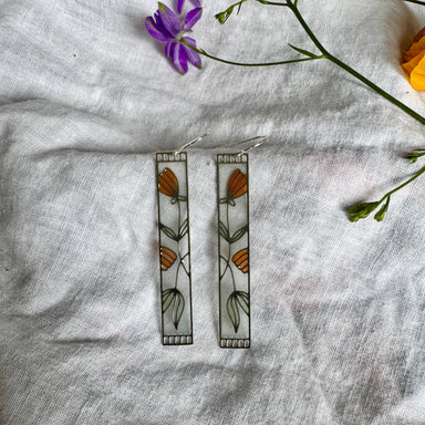 Honeysuckle Stained Glass Resin Earrings by Wild Lupine Folkcraft displayed on a textured white cloth with delicate flowers in the background, showcasing their modern botanical design and colorful resin.