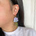 A model showcasing the Purple Iris earrings by Sunnie Creative, featuring a flower design with acrylic petals and leaves, worn on the ear for a stylish look.