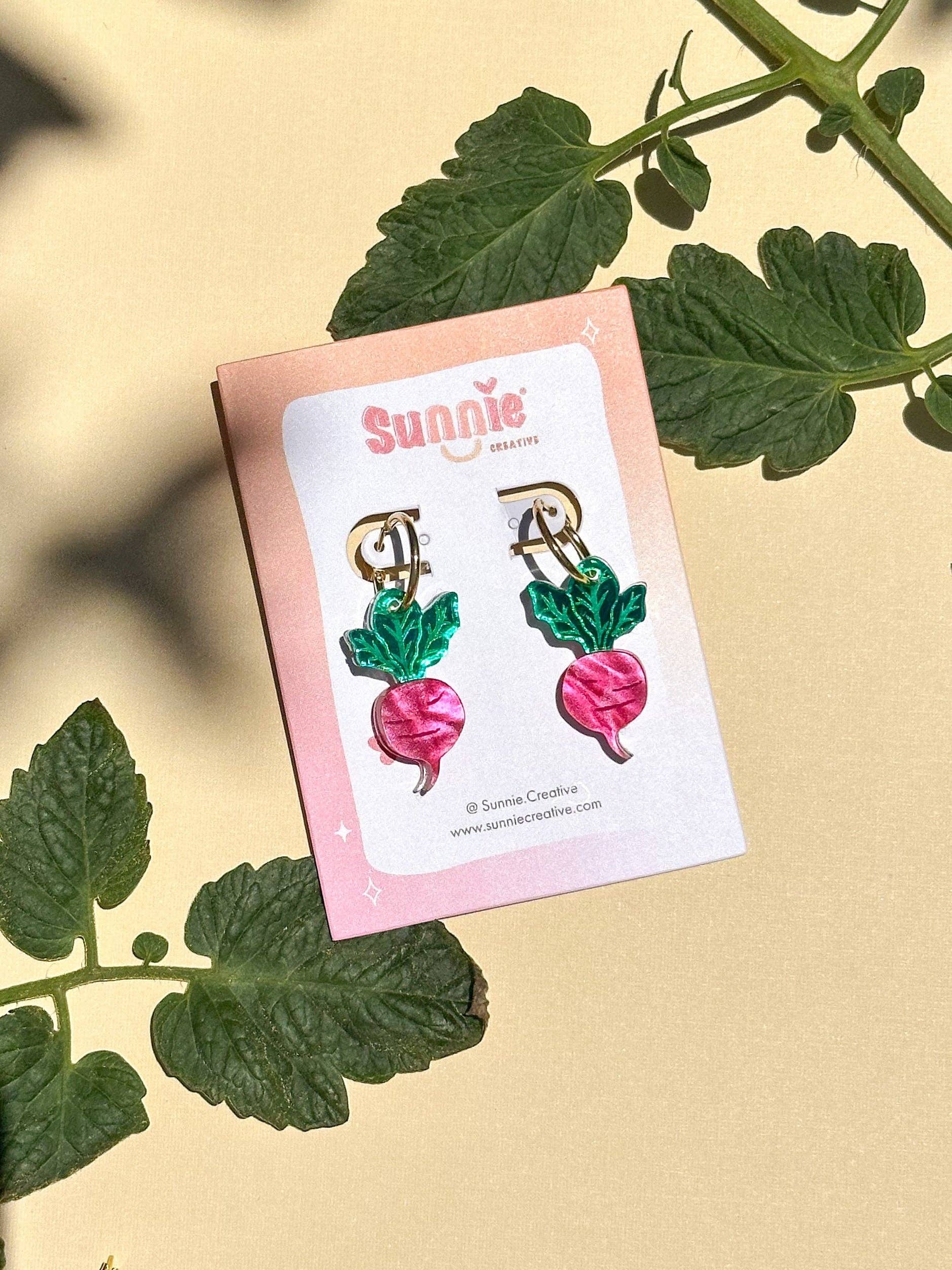 Mini Radish Dangle earrings displayed on a card by Sunnie Creative, featuring vibrant pink radish designs with green leaves, perfect for adding a playful touch to your outfit.