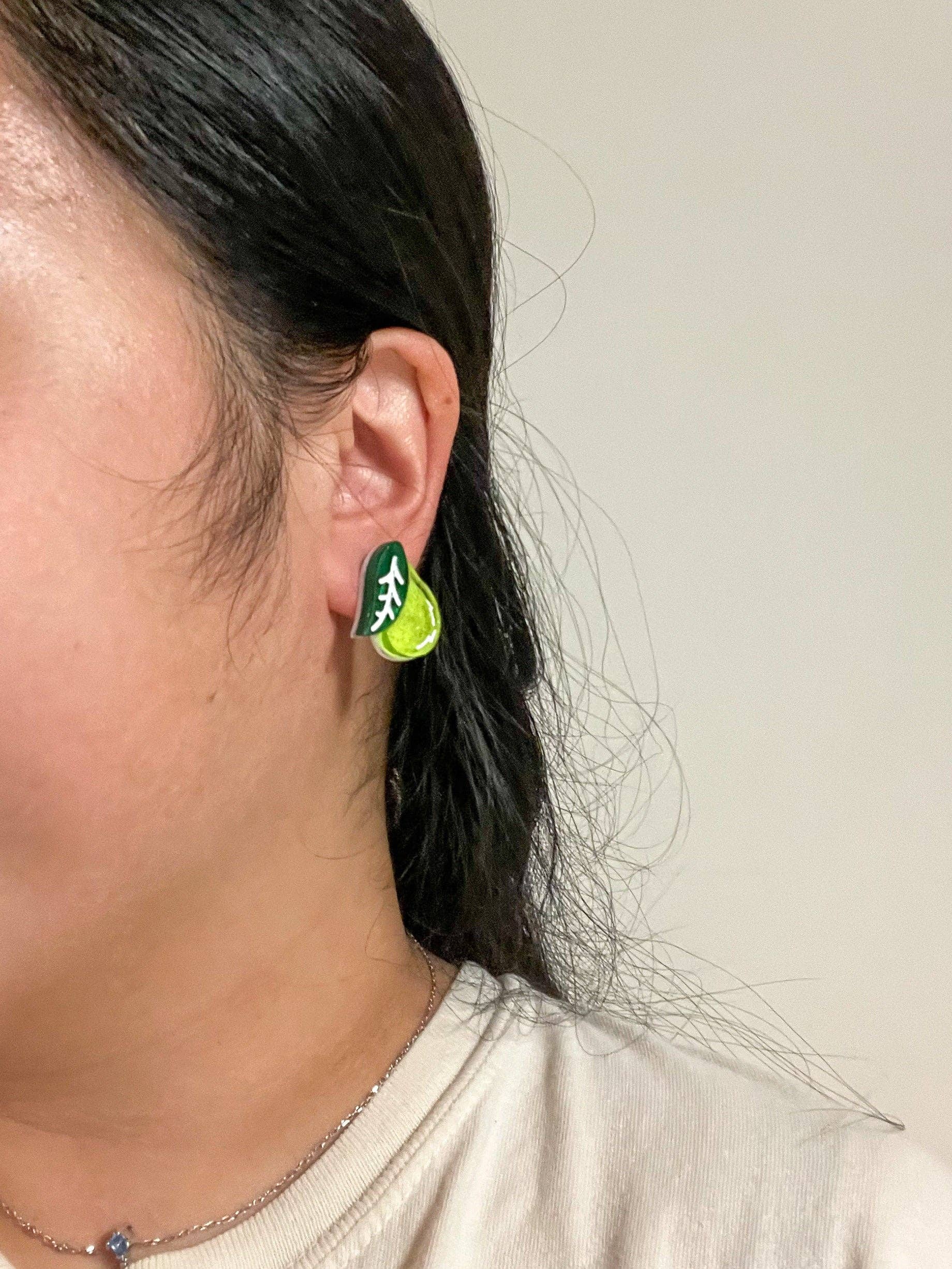 A close-up view of Mini Pear Dangle earrings by Sunnie Creative worn on a person's ear, illustrating the bright green acrylic pear design with leaf detail in a casual setting.