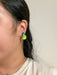 A close-up view of Mini Pear Dangle earrings by Sunnie Creative worn on a person's ear, illustrating the bright green acrylic pear design with leaf detail in a casual setting.