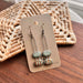 Turquoise Double Bubble Gold Striped Ceramic Earrings by The Artifact House displayed on a packaging card, featuring rustic woven decor in the background.