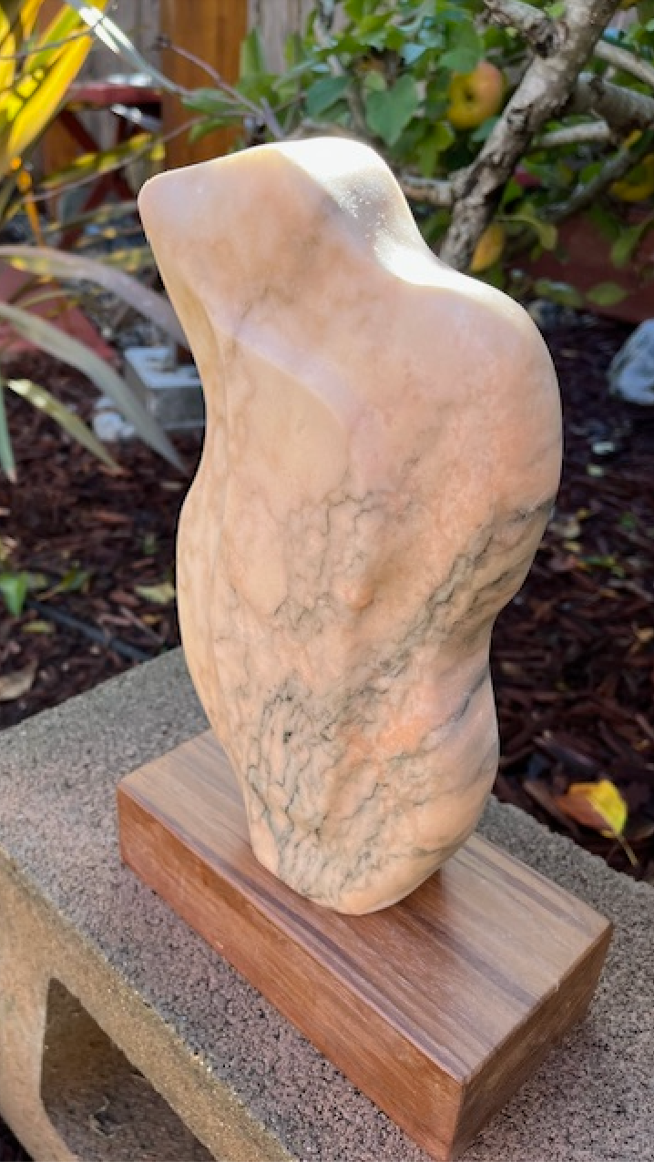 Hearts for the Arts | Stone Lover sculpture by Michael Bashista displayed on a wooden stand, showcasing a smooth, abstract form with a light beige and gray marble texture.