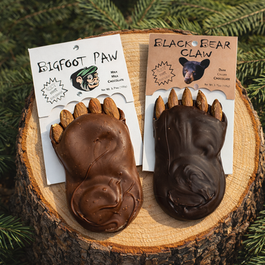 Two chocolate treats from Spokandy Chocolatier, featuring a Bigfoot Paw in milk chocolate with almonds and a Black Bear Claw in dark chocolate, elegantly displayed on a wooden log background, embodying the playful indulgence of Paw & Claw Chocolate.