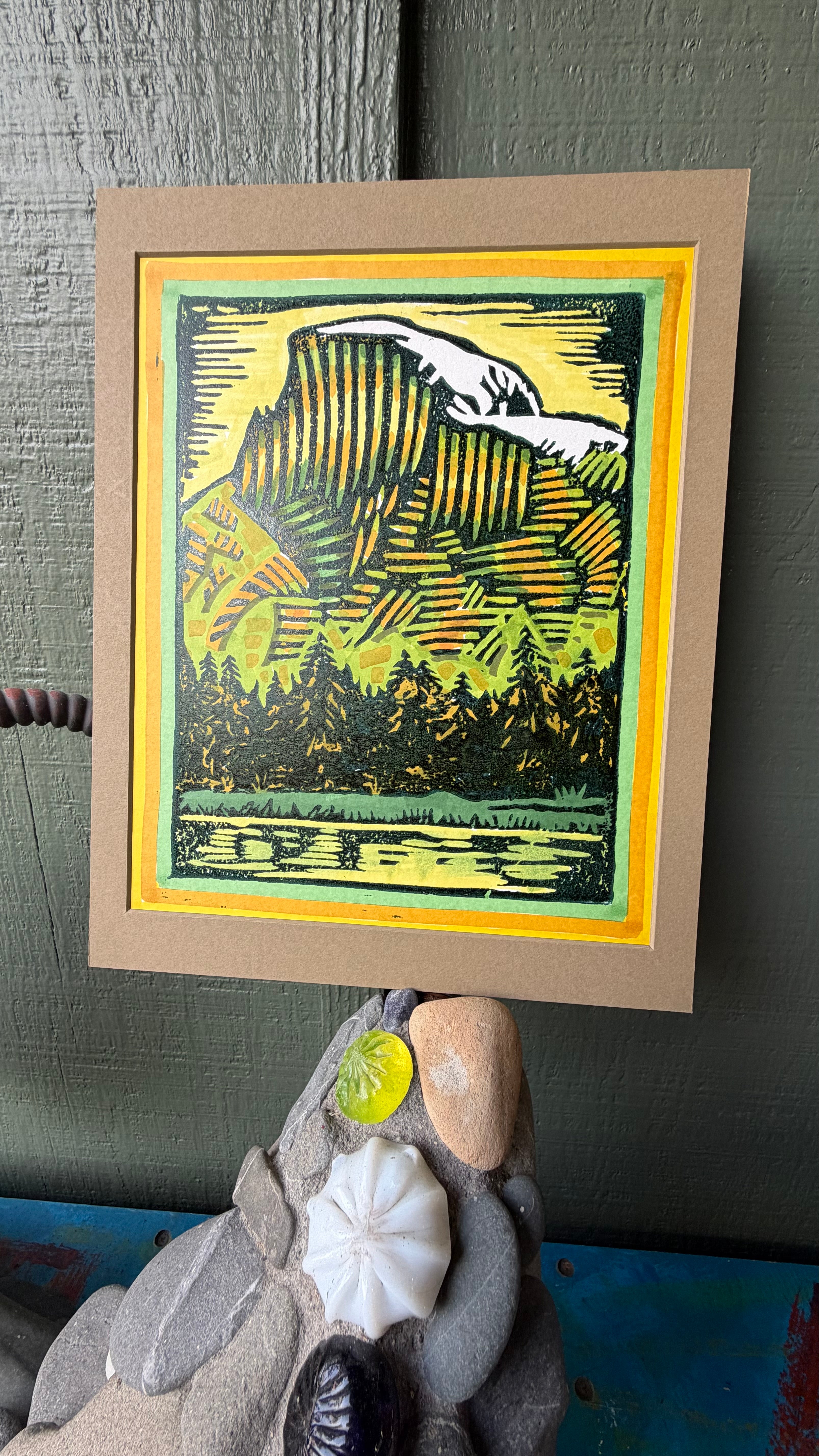 A vibrant linoleum block print titled 'Half Dome 1' by Hearts for the Arts, showcasing a yellow and green mountainous landscape with trees and water, framed with a decorative mat.